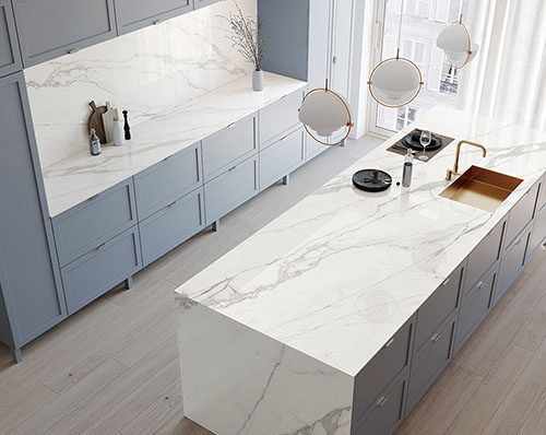 | R&D Marble, Inc. A modern white kitchen with porcelain countertops, backsplash, and island with light shining through the window.