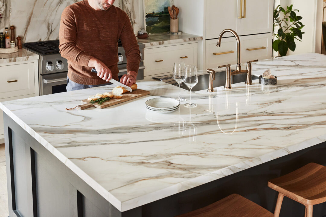 What are Upscale Kitchen Countertops & What Are Their Value? A man in a luxurious kitchen with upscale kitchen countertops slicing bread on a wood cutting board atop a white and gold porcelain surface.