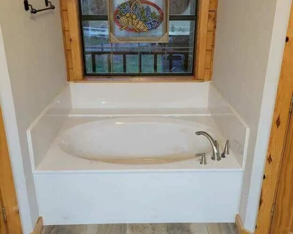 | R&D Marble, Inc. White built-in bathtub with surround and a curved faucet, window with wood trim behind featuring a stained-glass panel.