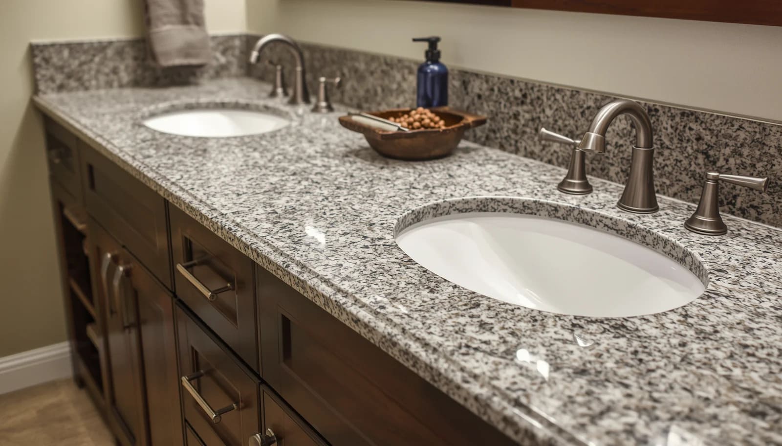 What Are Cultured Granite Countertops_ Cast Polymer Products Granite countertop double-sink vanity with bronze faucets, wooden cabinets, and a bowl of nuts beside a soap dispenser in a bathroom.
