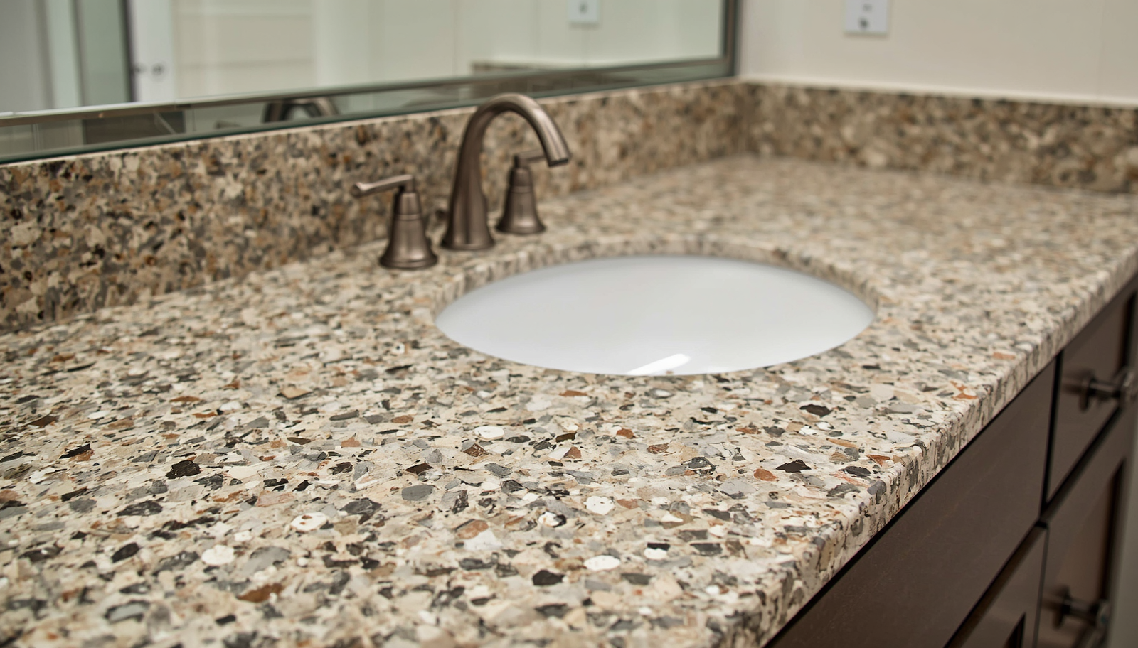 width_1600 1 | R&D Marble, Inc. Close-up of a speckled granite bathroom countertop with an oval undermount sink and bronze faucet.