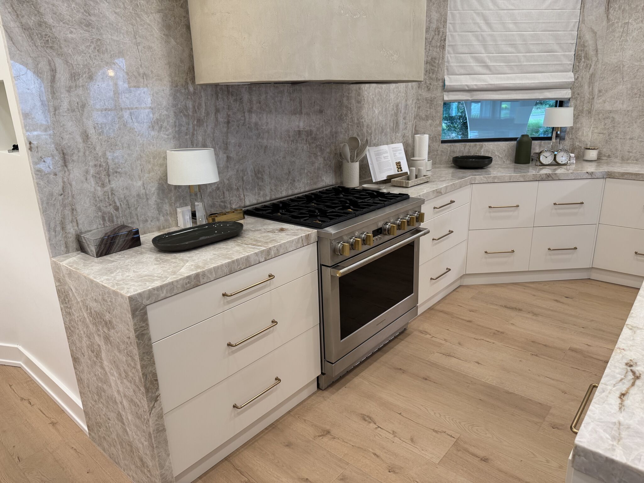 | R&D Marble, Inc. Modern white kitchen with marble countertops, stainless steel range, and brass hardware on drawers and cabinets.