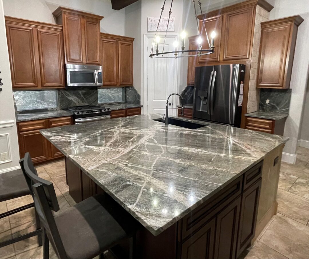 | R&D Marble, Inc. Kitchen with gray marble island, dark wood cabinets, stainless steel refrigerator and microwave, and a circular chandelier overhead.