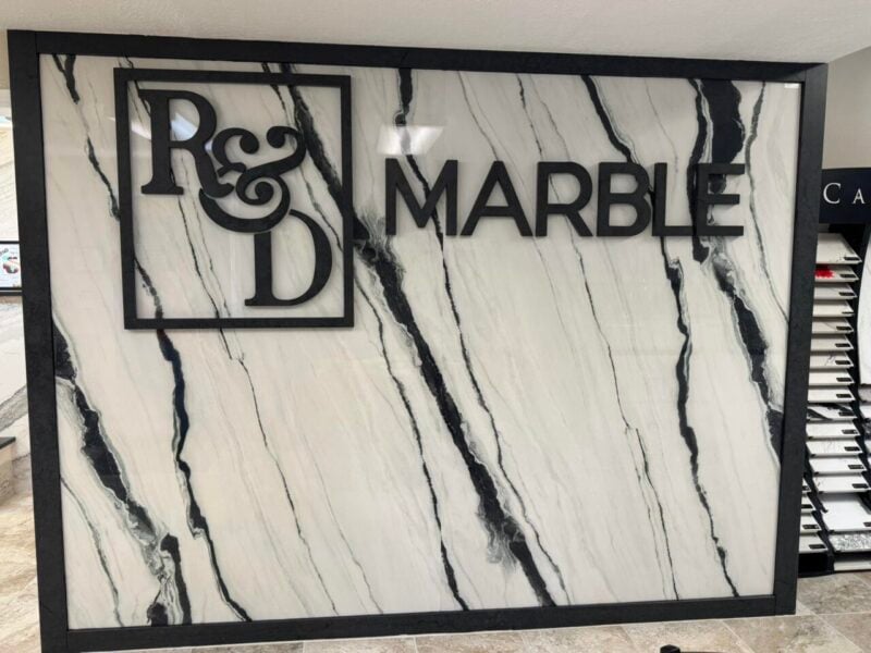 | R&D Marble, Inc. Framed sign reading R&D MARBLE on a white marble wall with black veins.
