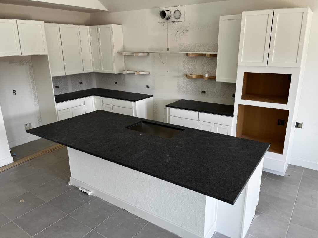 | R&D Marble, Inc. White kitchen with dark granite countertops, upper and lower cabinets, and an island under construction.