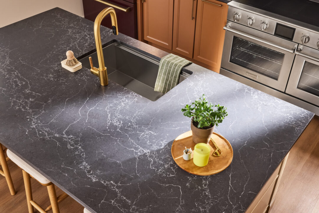 Popular Colors of Countertops for Kitchens in 2026 Modern kitchen island with a dark marble countertop, gold faucet, and gray sink beneath warm wood cabinets; stainless steel oven in the background and a small potted plant nearby.
