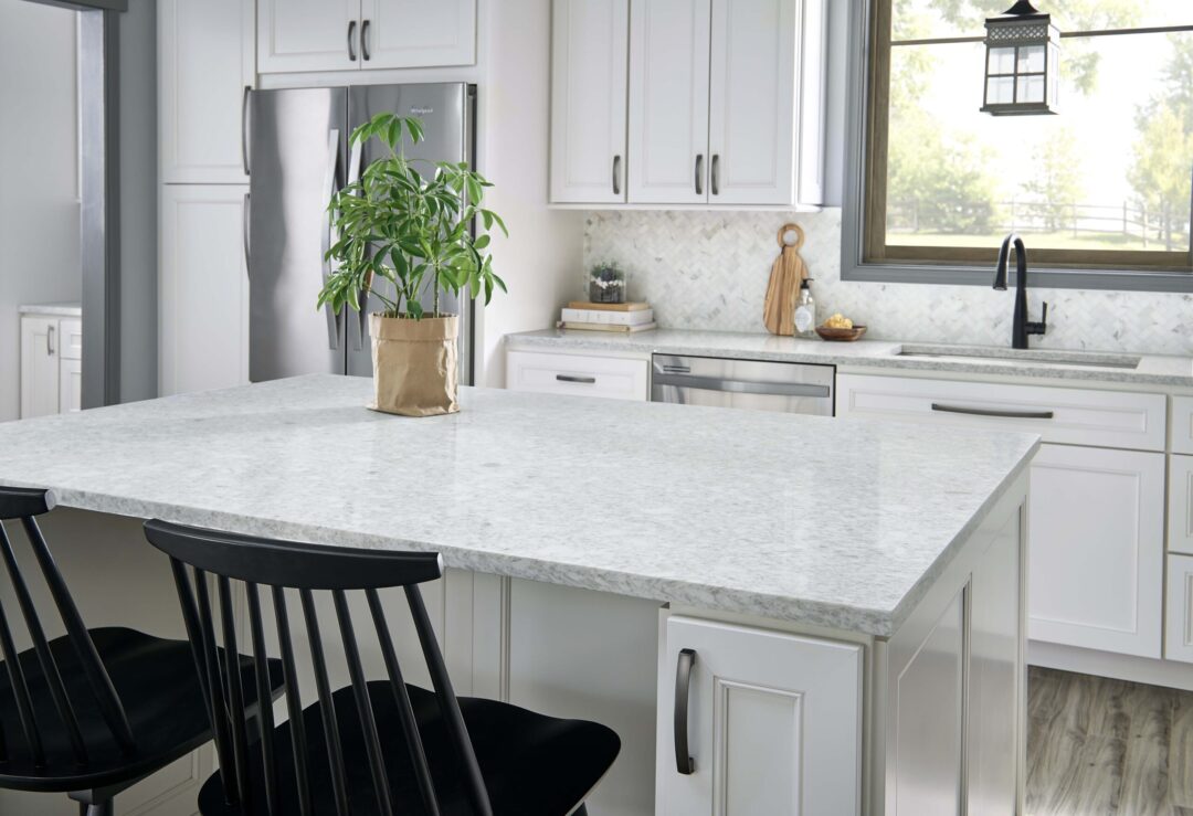| R&D Marble, Inc. Modern white kitchen featuring a marble island with a potted plant in a brown paper bag, black bar stools, and stainless steel appliances by a large window.