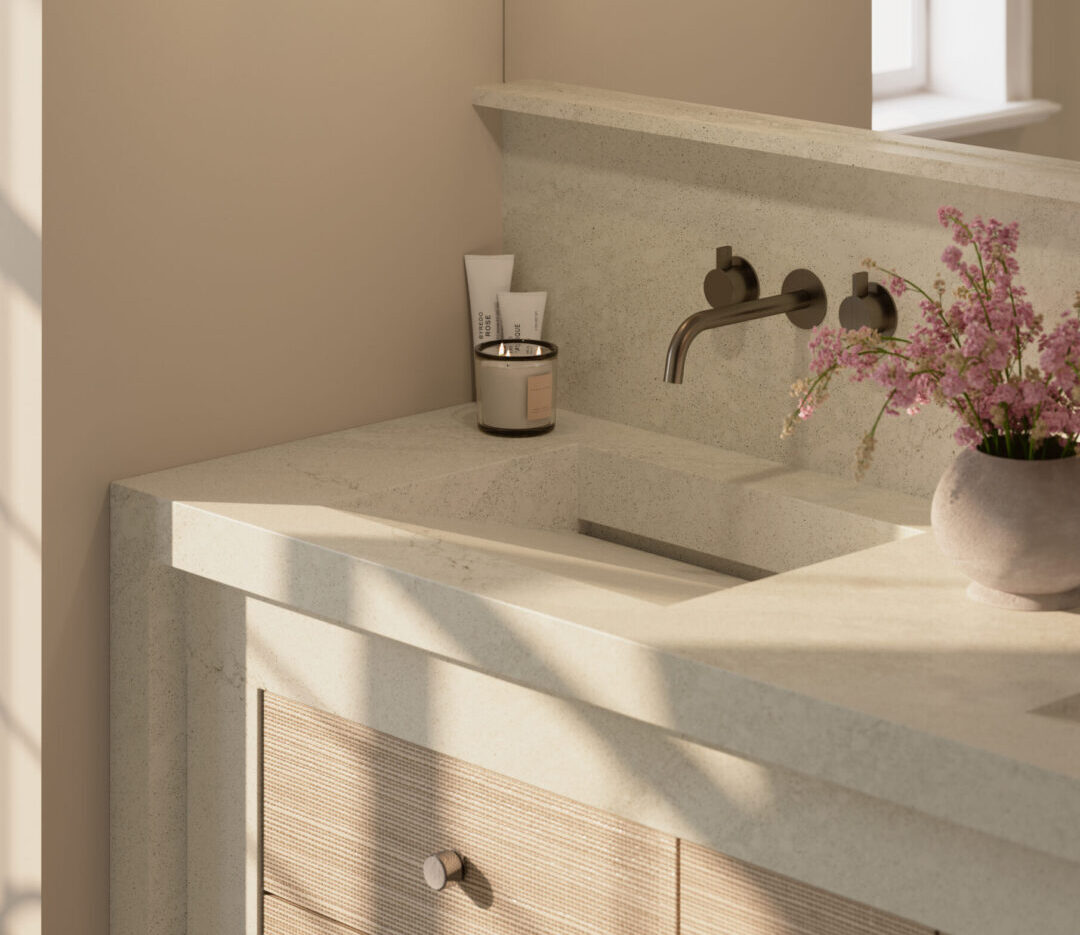 | R&D Marble, Inc. Beige bathroom vanity with a speckled stone countertop, wall-mounted faucet, and pink flowers in a vase.