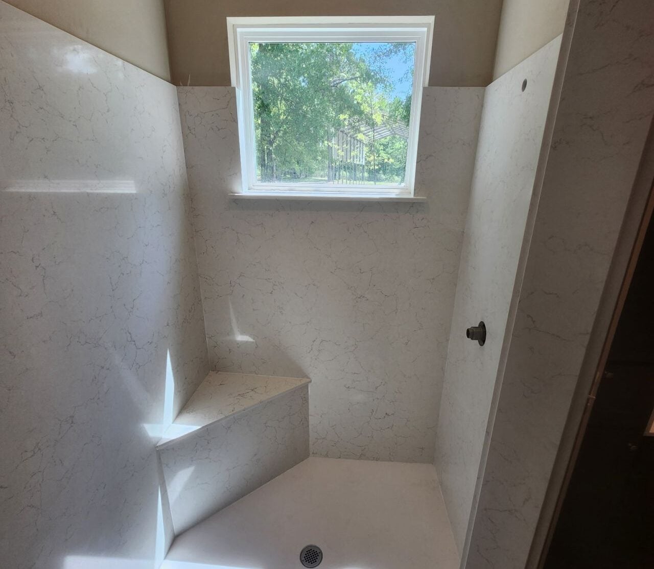 How to Upgrade Your Shower Walls with Beautiful Stone Shower interior with white marble walls, a small window showing greenery, and a triangular built-in bench.