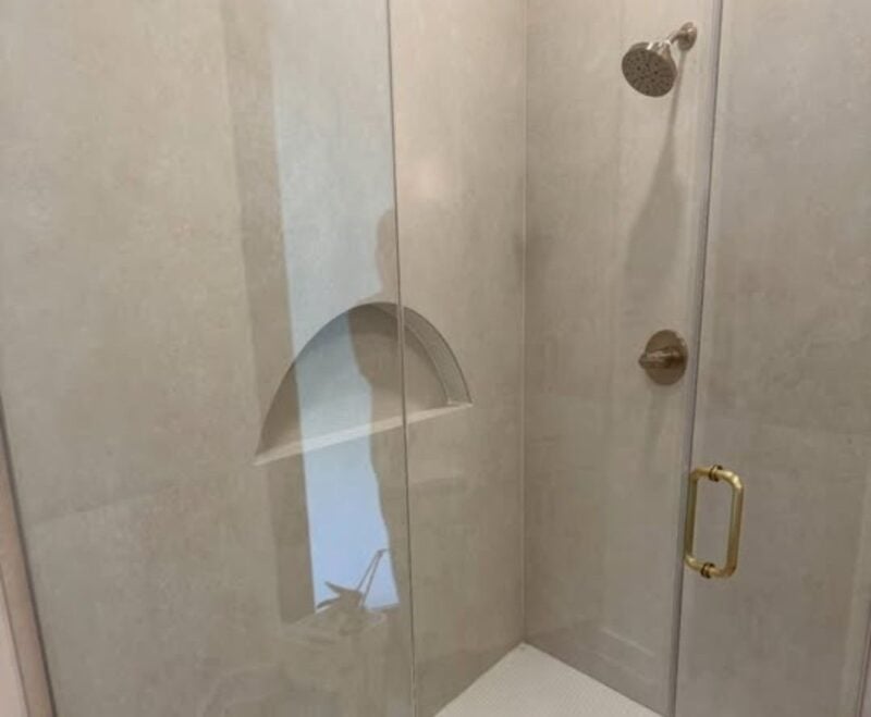 | R&D Marble, Inc. Glass-enclosed shower with a metal door handle and a rainfall showerhead, light gray tiled walls, and a square drain on a white tiled floor.