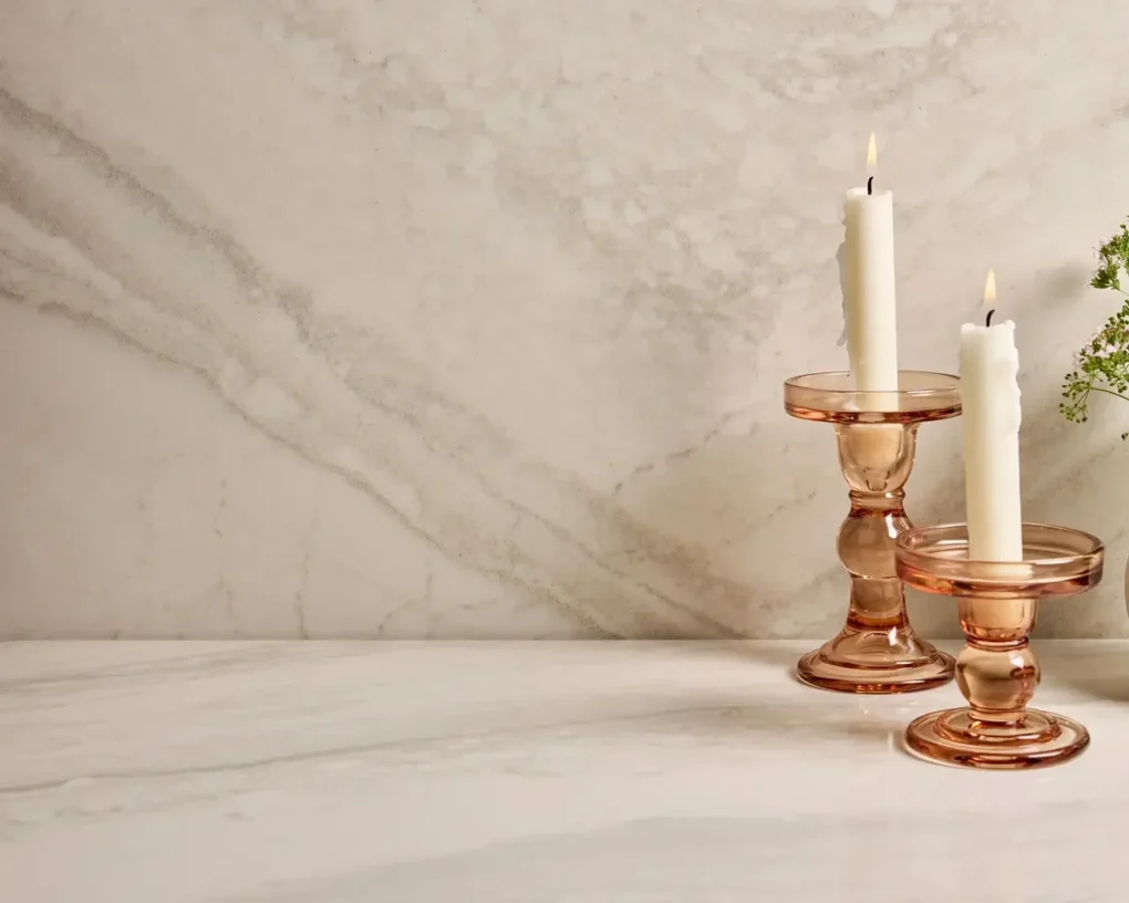 Two tall white candles in pink glass candle holders on a marble tabletop, with a soft background and a small sprig of greenery nearby.