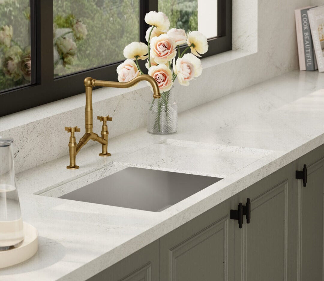Brass kitchen faucet with white speckled countertop, vases of pale pink flowers by a large window overlooking hills.
