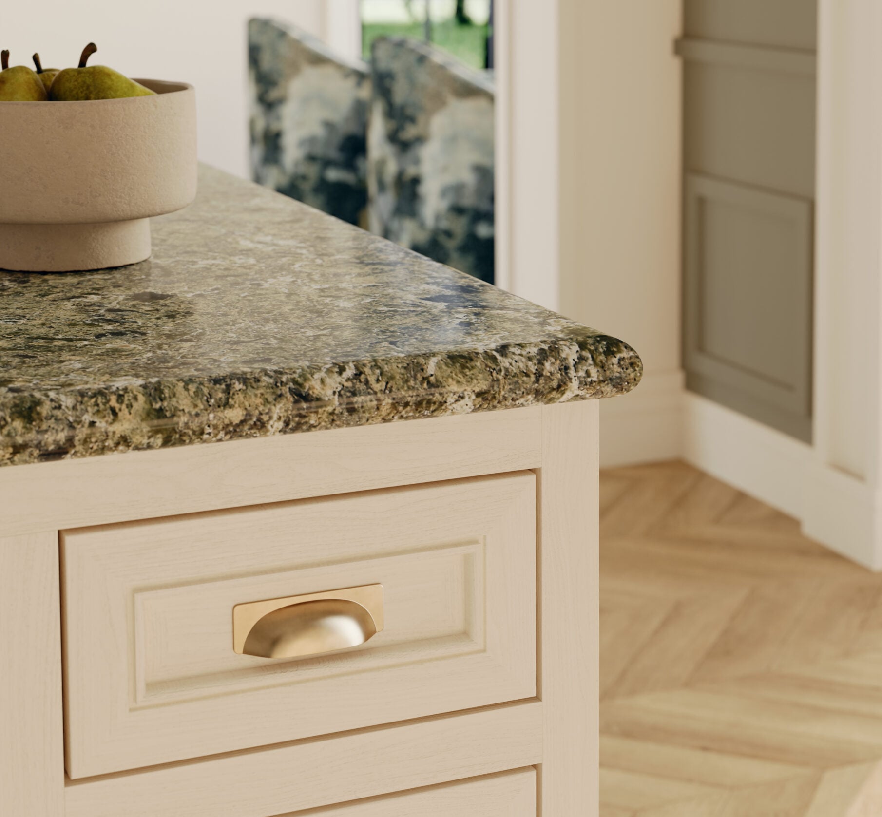 Cream-painted kitchen island with a speckled granite countertop, a beige bowl of pears, and brass drawer pulls in the foreground.