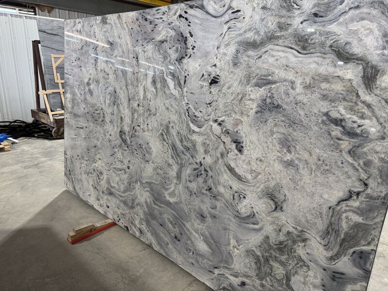 | R&D Marble, Inc. Large polished stone slab with swirling gray and white veining leaning against other slabs in a warehouse.