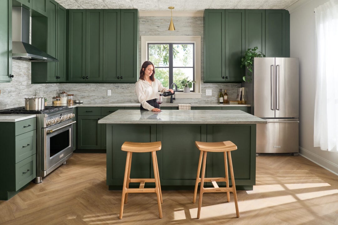 Woman pouring red wine into a glass at a marble kitchen island, with dark green cabinets and stainless steel fridge nearby.