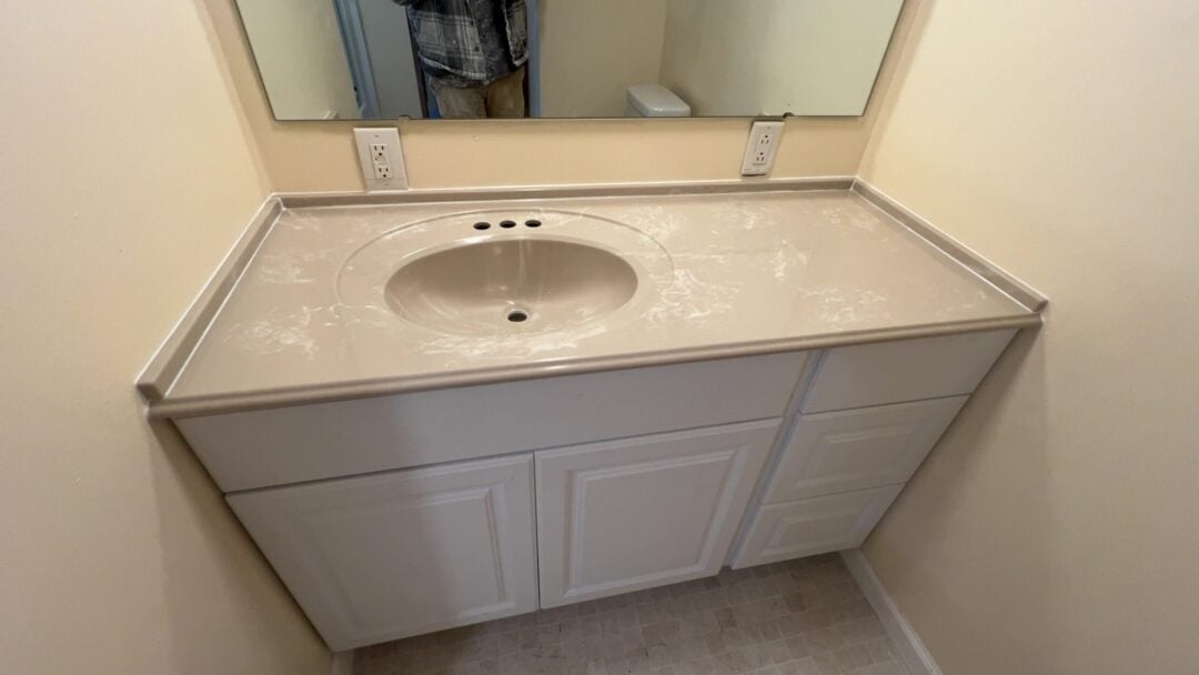 | R&D Marble, Inc. Bathroom vanity with light gray cabinets, an oval sink cutout, and faucet holes; mirror and outlets on the beige wall.