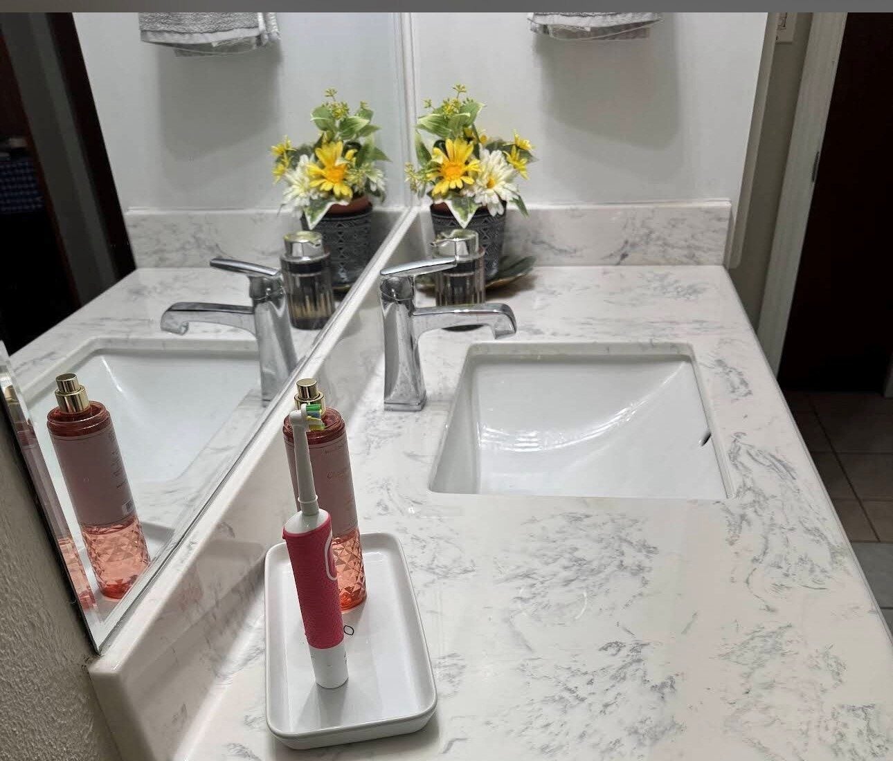 How to Clean Engineered Marble Vanity Top: Complete Guide White marble bathroom counter with two sinks and a chrome faucet, mirror, and yellow flower arrangements in vases; a pink electric toothbrush and pink bottle on a small tray.