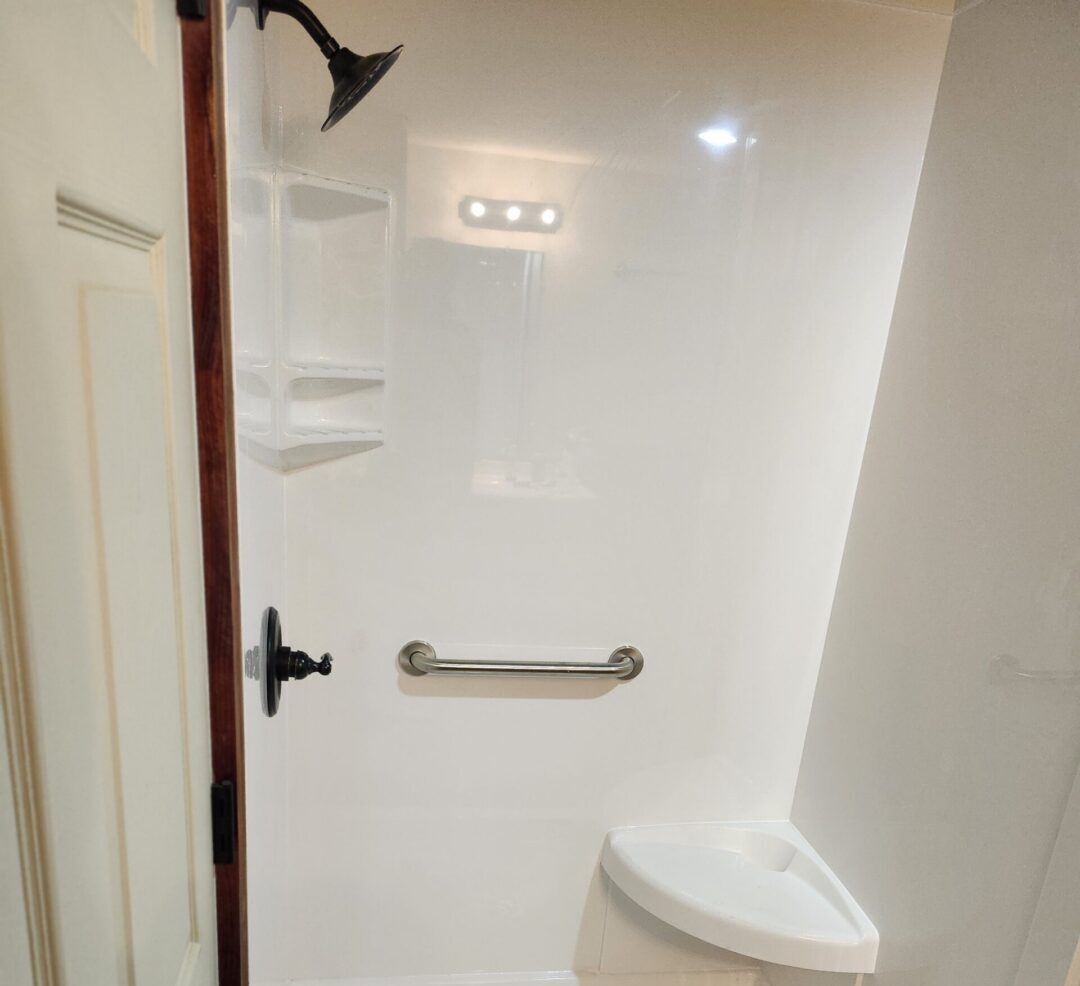 Walk-in shower with white acrylic walls, a chrome grab bar, and a small built-in shelf near the corner.