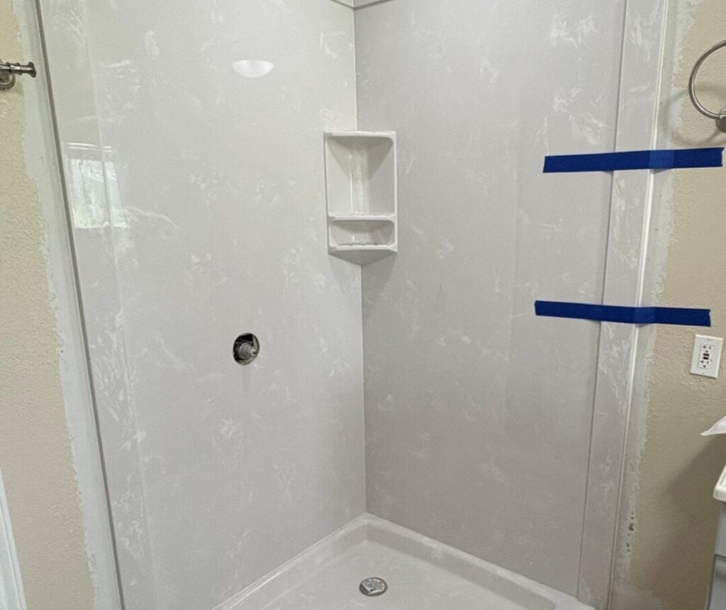 Cultured Marble Shower Installed by R&D Marble | R&D Marble, Inc. White corner shower with built-in niche, blue painter's tape on the walls, and an unfinished plywood floor (under construction).