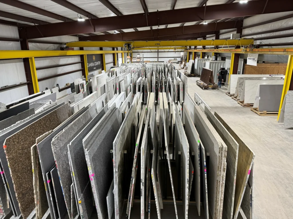 Warehouse interior with tall racks of vertical stone slabs arranged in aisles and a yellow overhead crane crossing the ceiling, used for storing and moving heavy countertops and tiles.