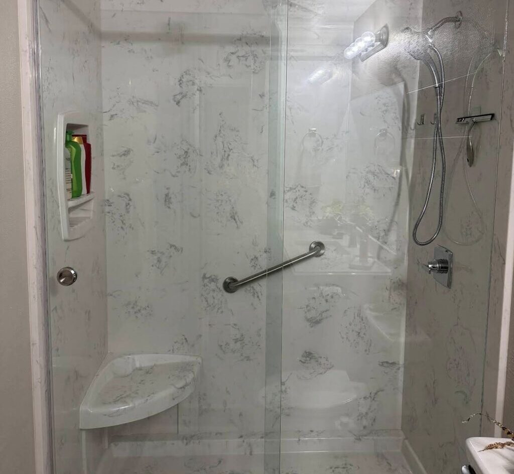 White Cultured Marble Shower Installed by R&D Marble | R&D Marble, Inc. Glass-enclosed shower with marble walls, sliding door, and chrome hardware; handheld shower, built-in corner shelf with toiletries, and a small bench inside.