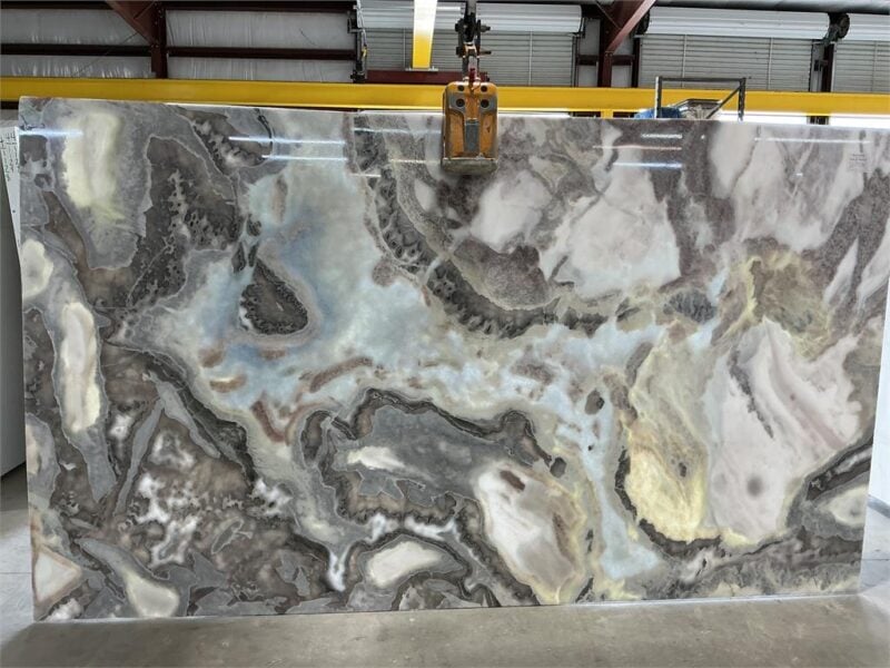 Large polished marble slab with gray, white, and beige veining suspended by a yellow lifting clamp in an industrial warehouse.