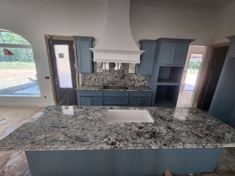 Under‑construction kitchen with blue cabinets, granite countertops, and a white range hood over a cooktop; large island with an undermount sink.
