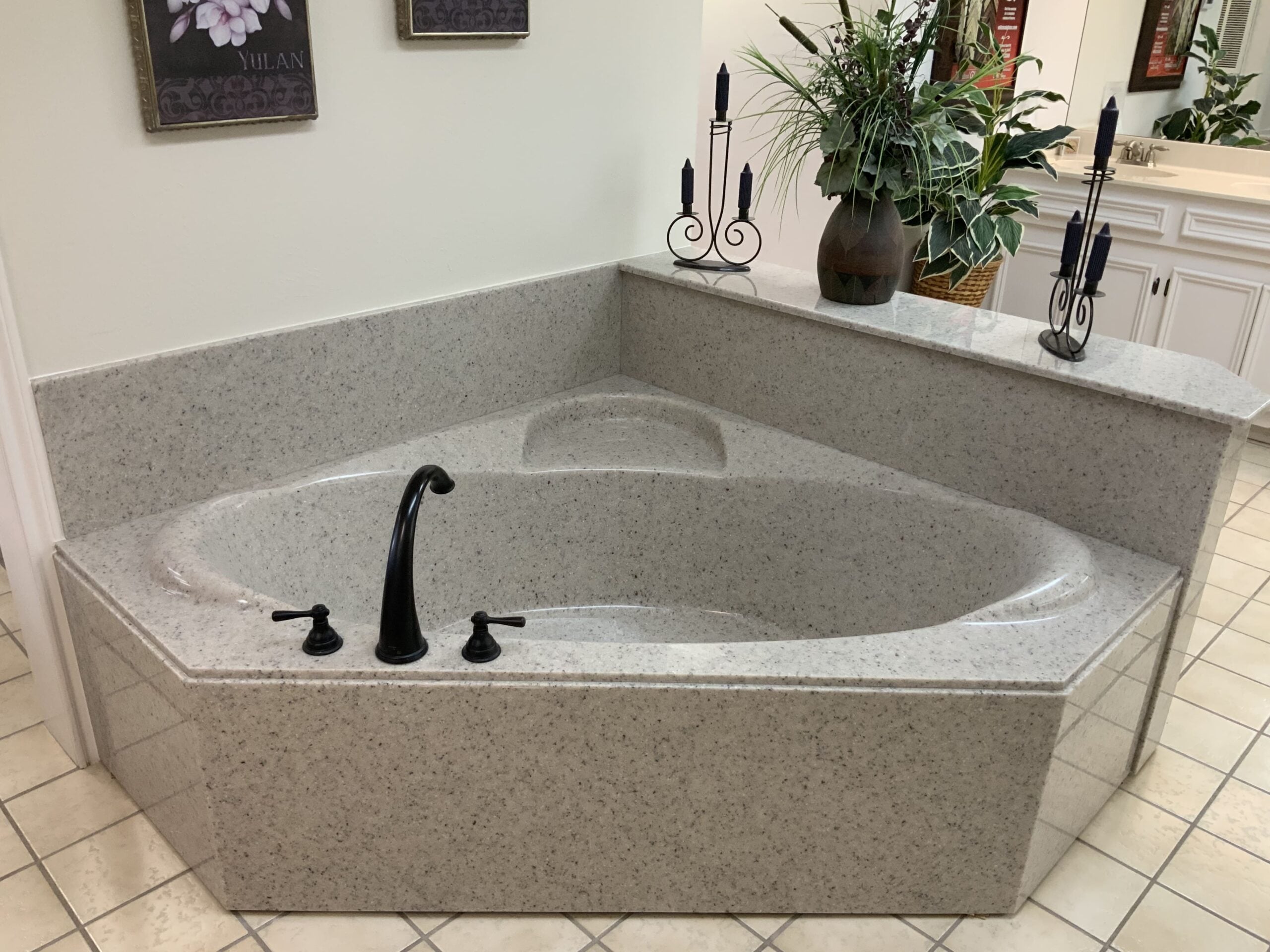 Cultured-granite-1 | R&D Marble, Inc. Stone-gray granite bathtub with curved edge and black faucet, set in a bathroom alcove with candle holders and potted plants on the ledge; white cabinetry visible in the background.