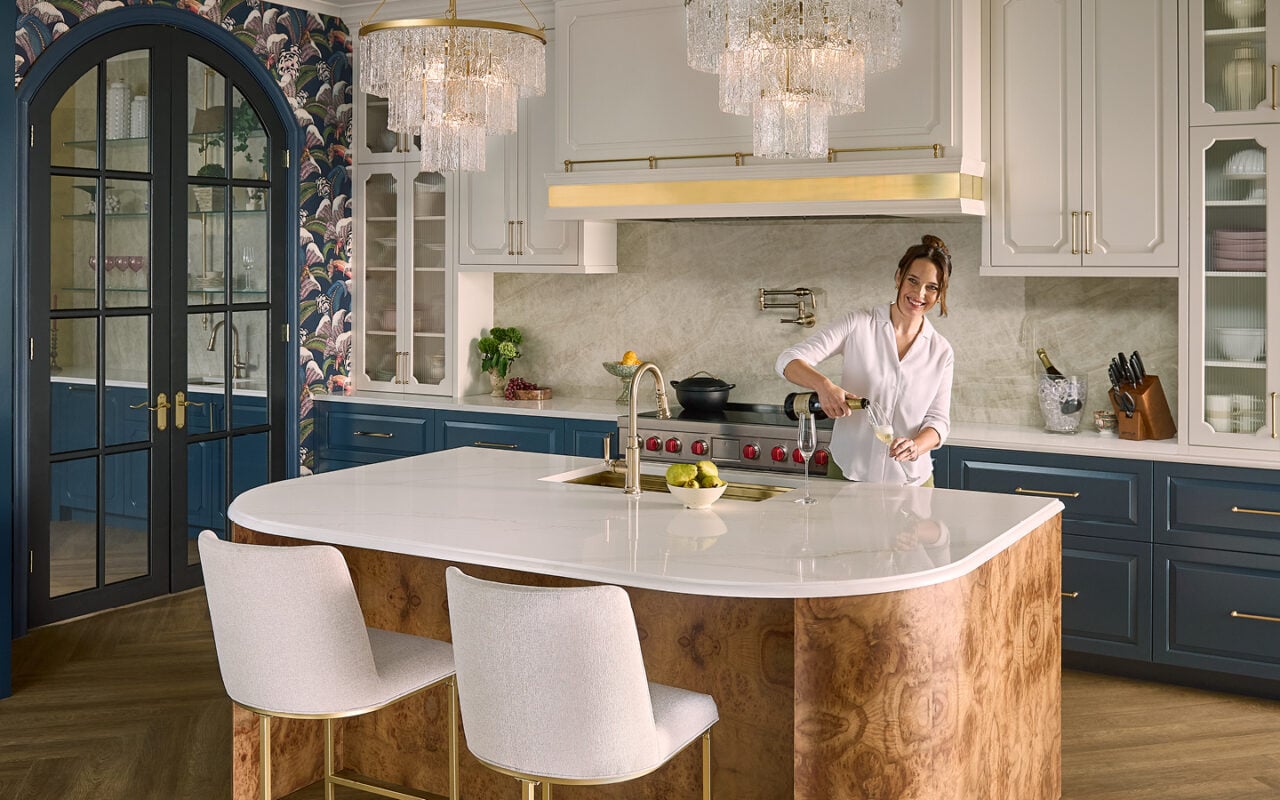 A woman pours champagne into a flute at a large marble-topped kitchen island in a luxe white-and-navy kitchen with crystal chandeliers.
